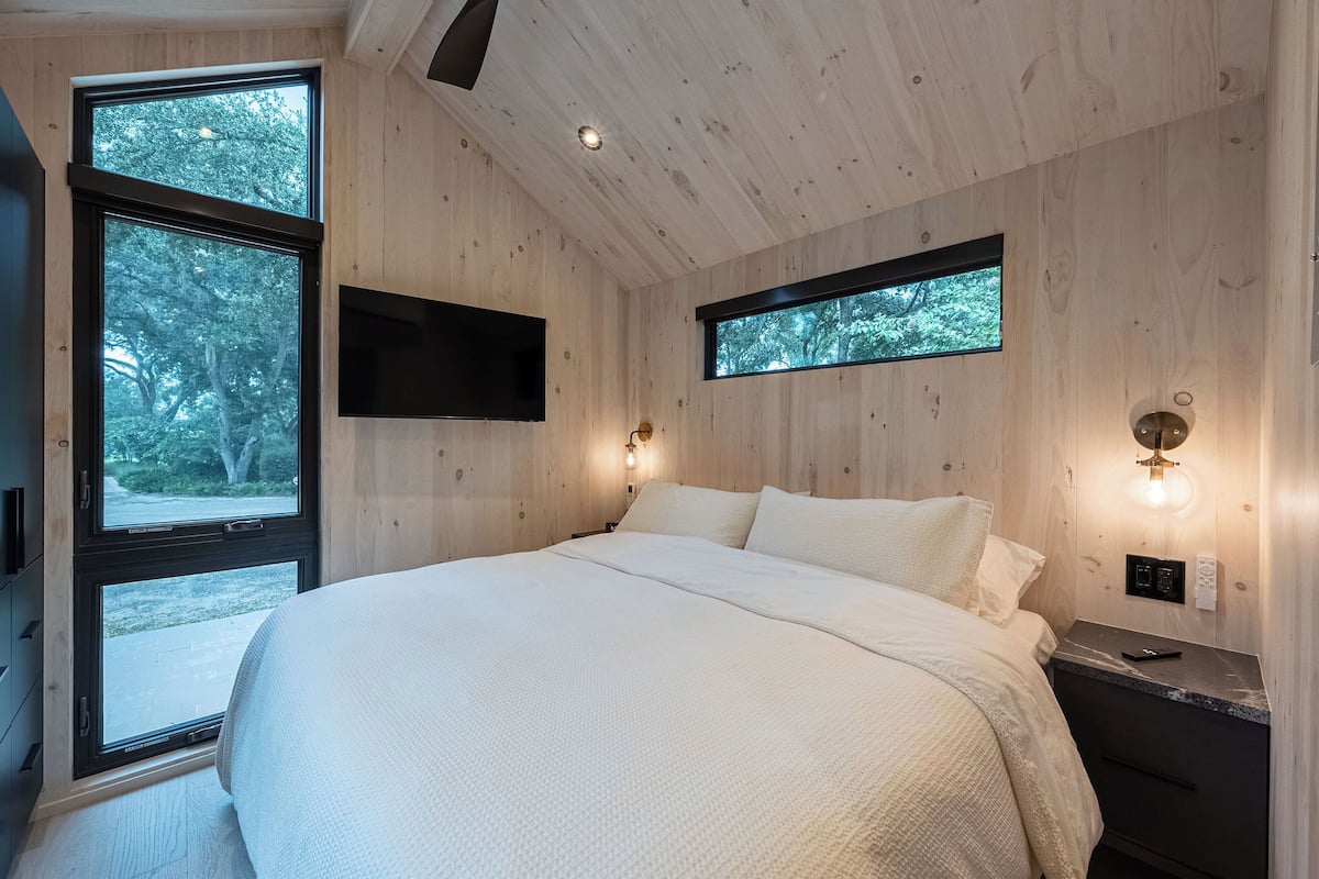 A bedroom with a large bed, wooden walls, a TV mounted on the wall, and two windows providing views of trees.