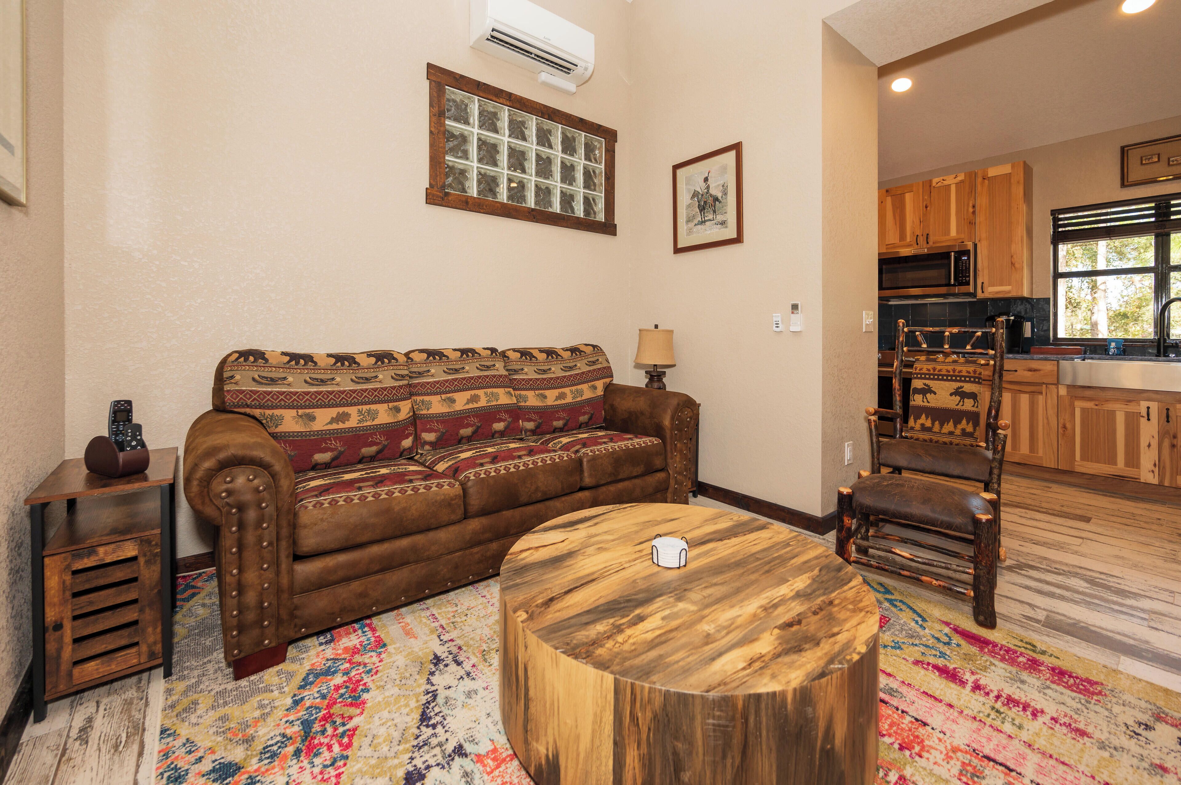 A living room with a brown sofa, a wooden coffee table, a patterned rug, and a kitchen in the background.