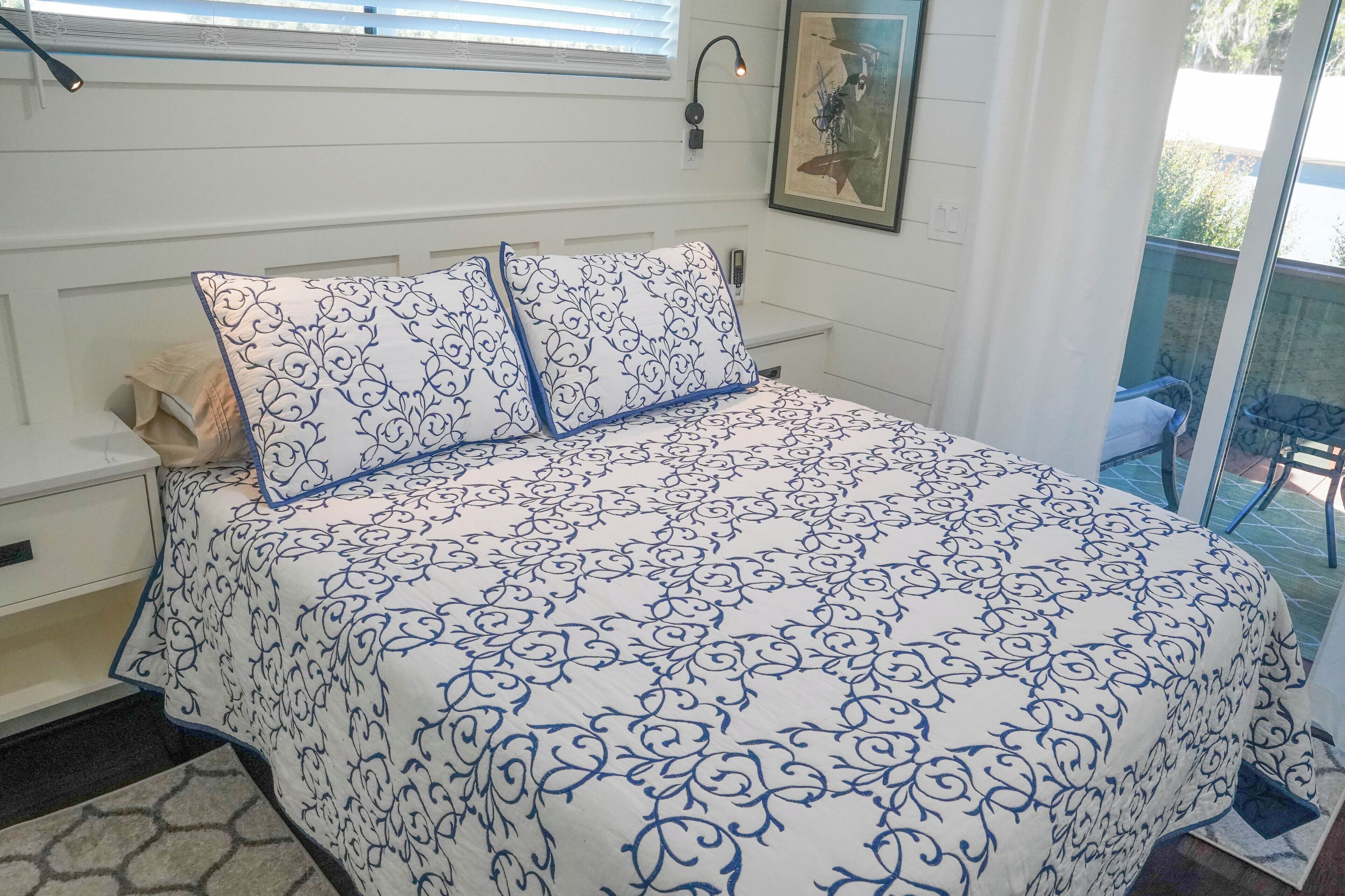A bedroom with a bed covered in a blue and white patterned bedspread, a nightstand with a lamp, and a framed picture on the wall.