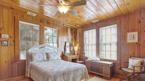 A wooden cabin bedroom with a bed, dresser, chair, and large windows.