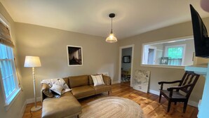 A living room with a brown leather sofa, a round woven rug, a wooden floor, a standing lamp, and a television mounted on the wall. - Charming Apt above Bookshop in Historic Tiverton Four Corners Village Center! (Tiverton)
