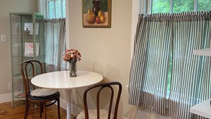 A dining area with a round table, two chairs, a vase with flowers, a painting, and a window with curtains. - Charming Apt above Bookshop in Historic Tiverton Four Corners Village Center! (Tiverton)