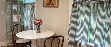 A dining area with a round table, two chairs, a vase with flowers, a painting, and a window with curtains.