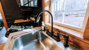 A kitchen with a stainless steel sink, a stove, and a window with a view of trees. - Charming cabin for 2, with private hot tub and peaceful creek (Beach City)