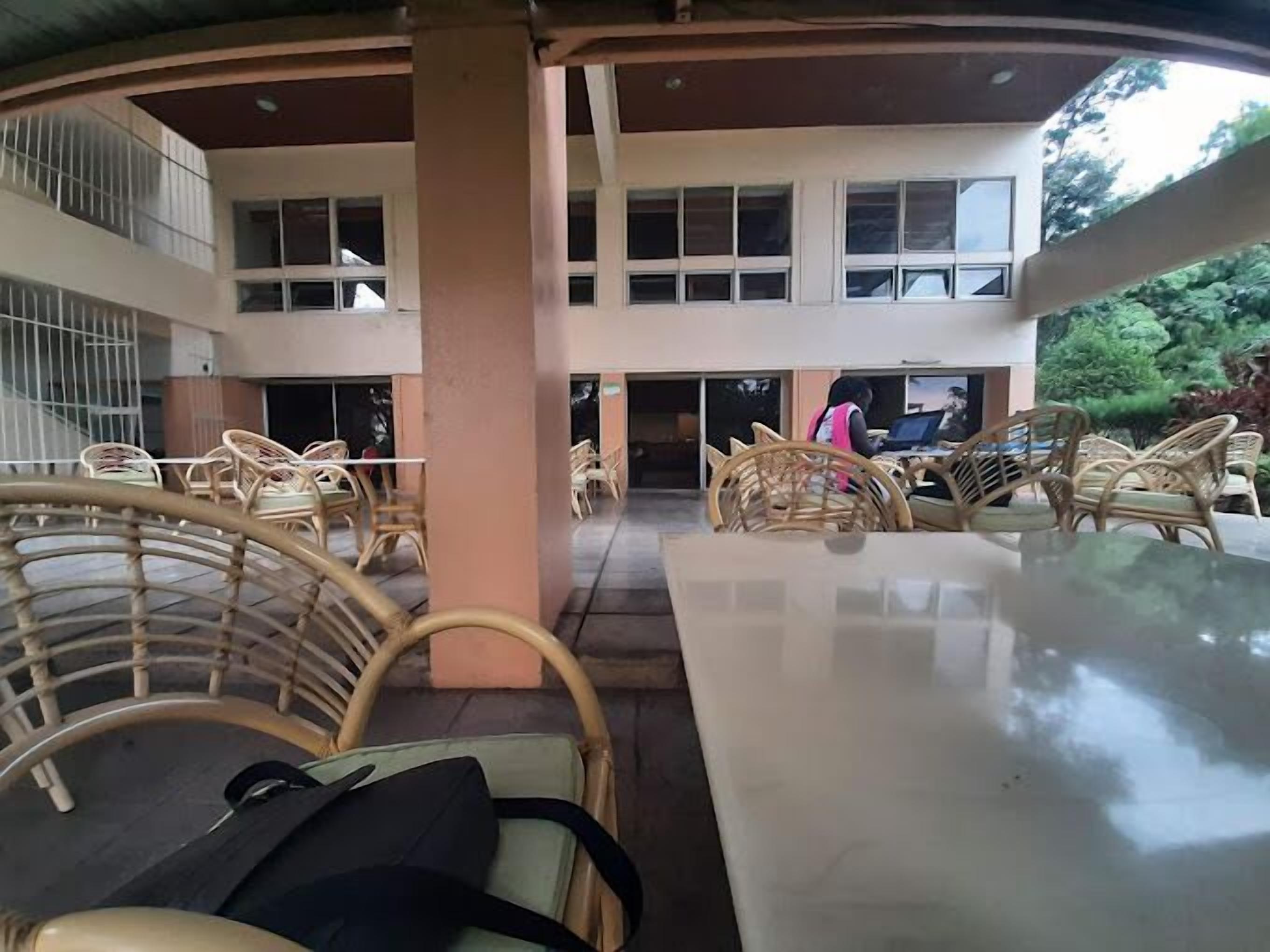 An indoor seating area with wicker chairs and tables, a central pillar, and a view of the building's exterior through large windows.