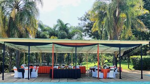 A covered outdoor dining area with tables and chairs, a pool in the foreground, and lush greenery in the background.
