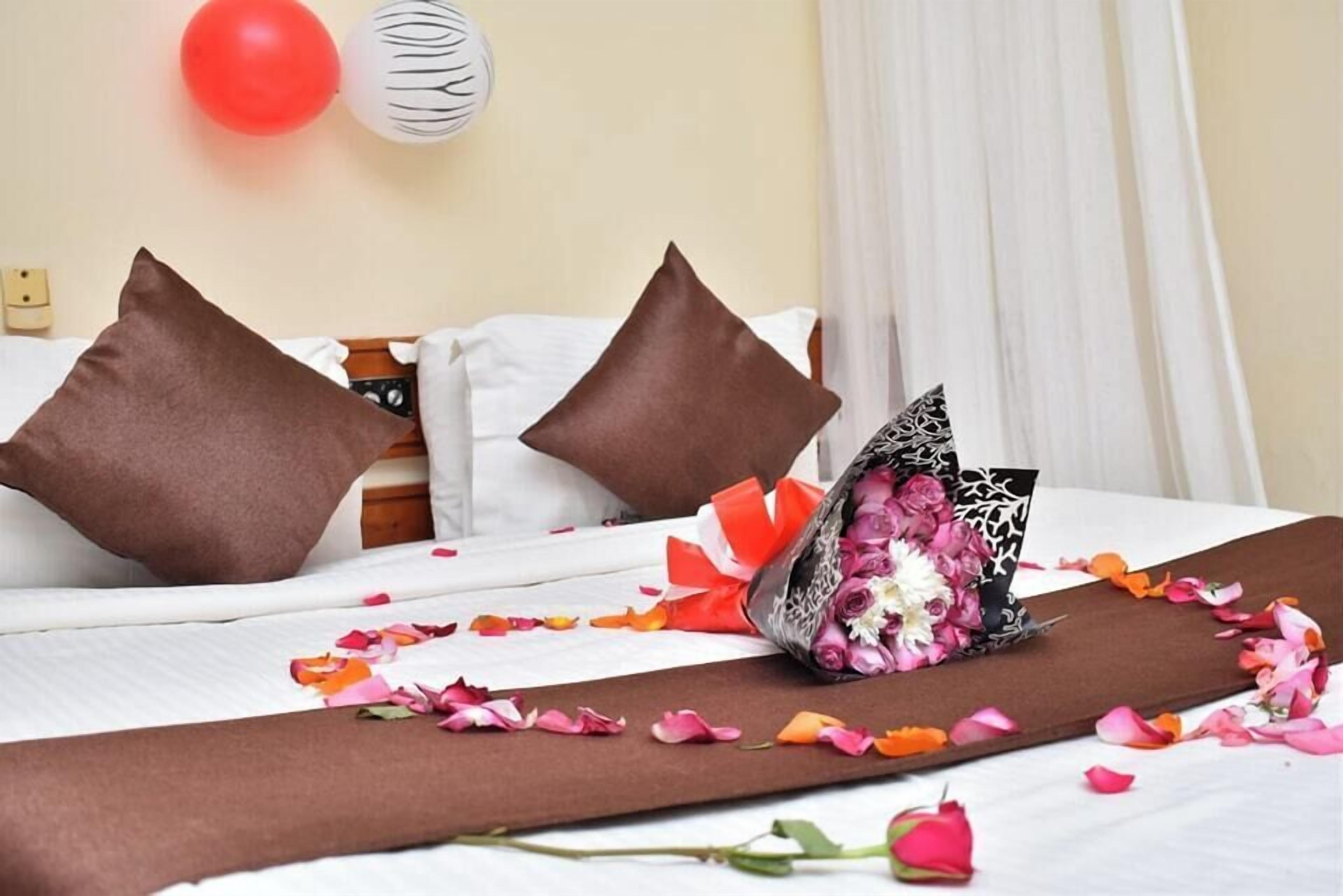Quarto de hotel com cama, almofadas, um buquê de flores e uma rosa sobre a cama.