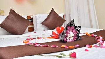 Quarto de hotel com cama, almofadas, um buquê de flores e uma rosa sobre a cama.