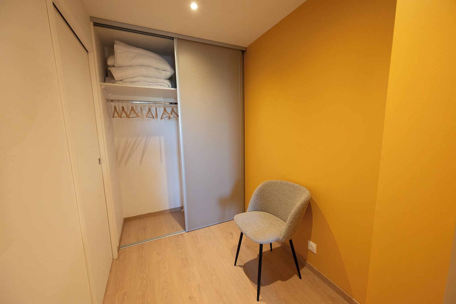 Een kamer met een houten vloer, een stoel, een kledingkast met hangende kleding en een gele muur.