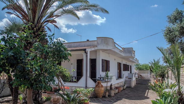 A white building with a tiled roof, surrounded by potted plants and palm trees. - Villa Ada (Leporano Marina)