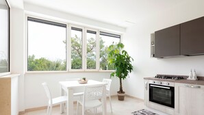 Une cuisine moderne avec des meubles blancs, une table à manger avec des chaises, une plante en pot et une grande fenêtre donnant sur des arbres.