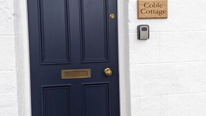 En mörkblå dörr med ett mässingshandtag och en mässingsplakett, en träskylt med texten ”Coble Cottage”, och en dörrklocka på väggen.
