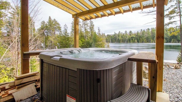 A hot tub under a wooden pergola with a view of a lake and forest. - Timber Wilderness Lodge (North Frontenac)