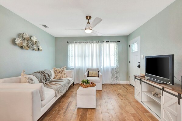 A living room with a white sofa, a white armchair, a TV on a wooden stand, and a ceiling fan.
