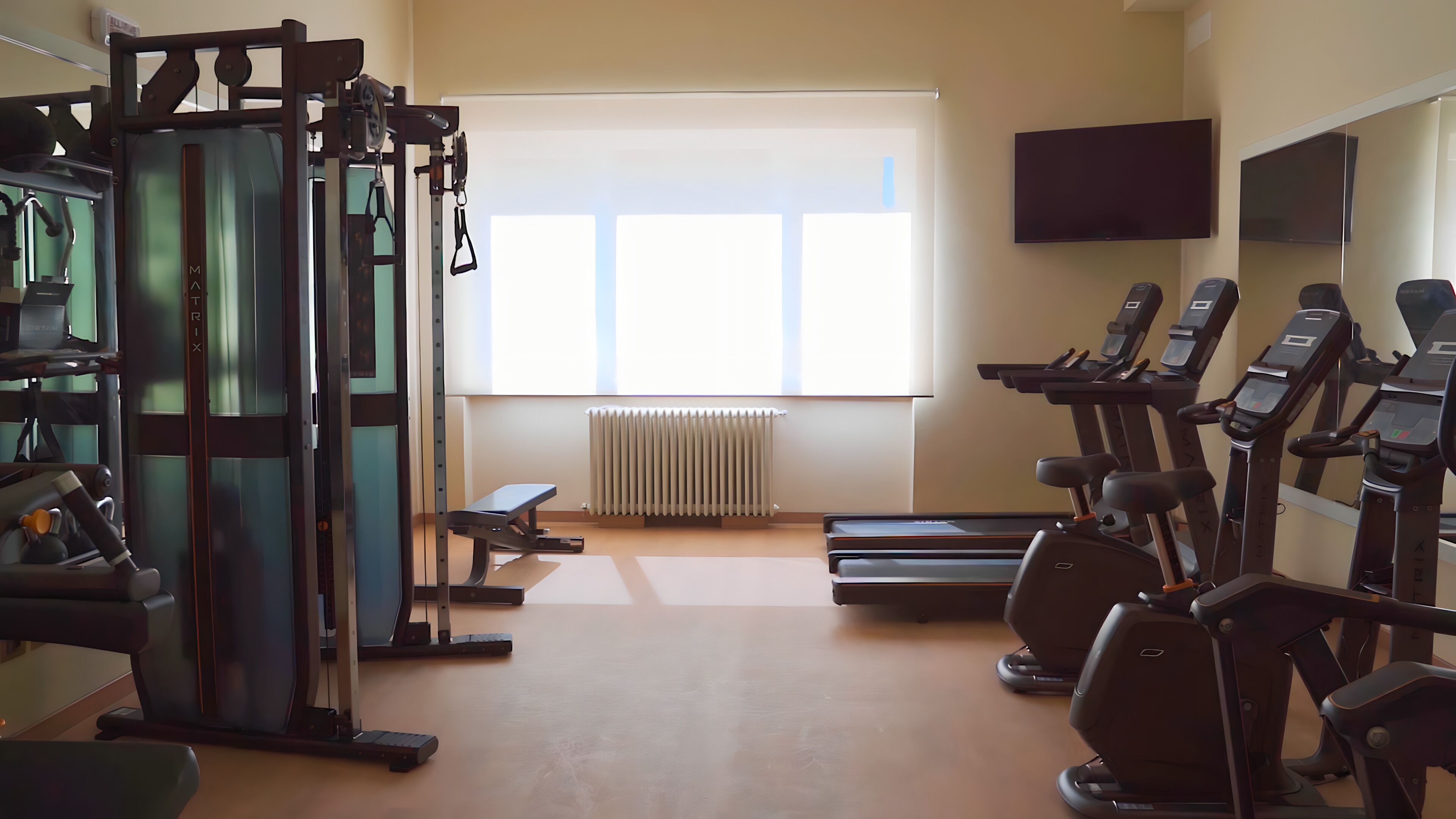 Ein Fitnessraum mit Trainingsgeräten, einer Bank, einem Laufband und einem Fernseher an der Wand.