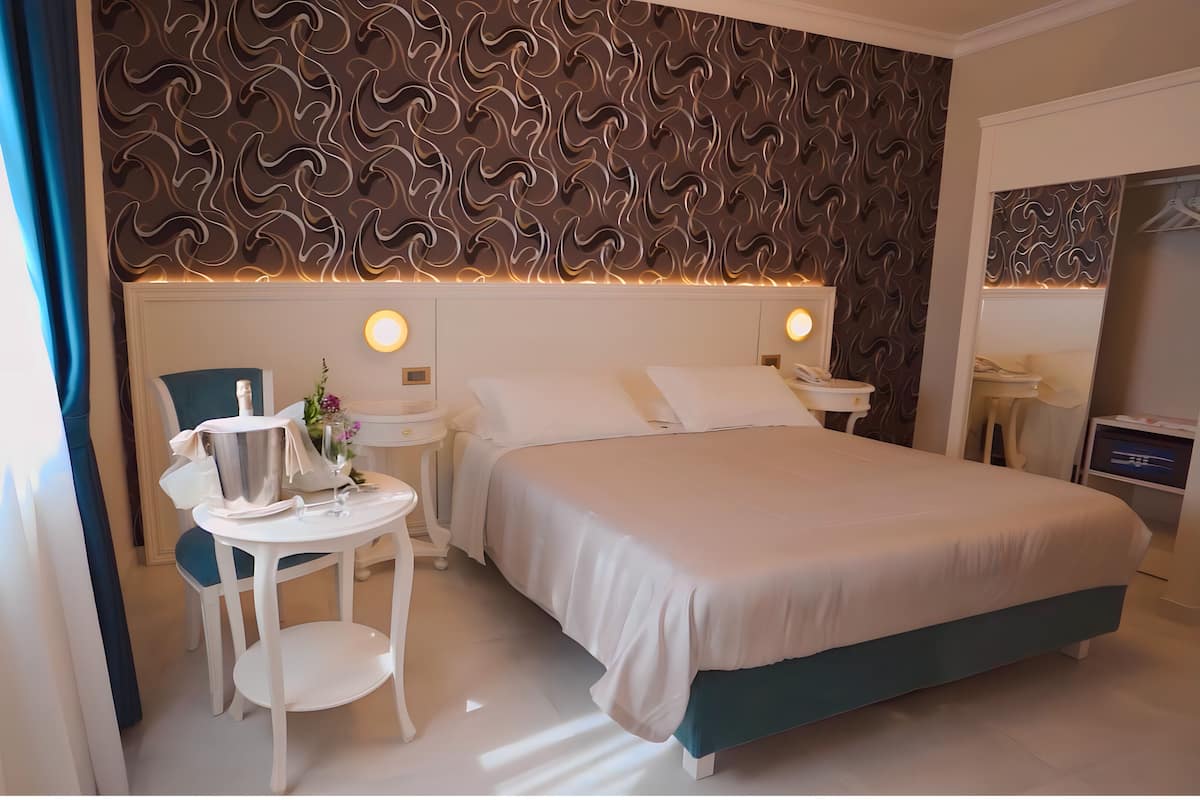 Una habitación de hotel con una cama grande, dos mesitas de noche, una pequeña mesa con una botella y copas, una silla y un papel tapiz estampado.