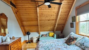 A cozy bedroom with a sloped ceiling, wooden beams, a bed with floral bedding, a nightstand, a dresser, and a window with a view of trees.
