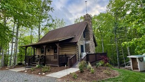 A rustic cabin with a stone chimney, wooden porch, and a small shed in a wooded area.