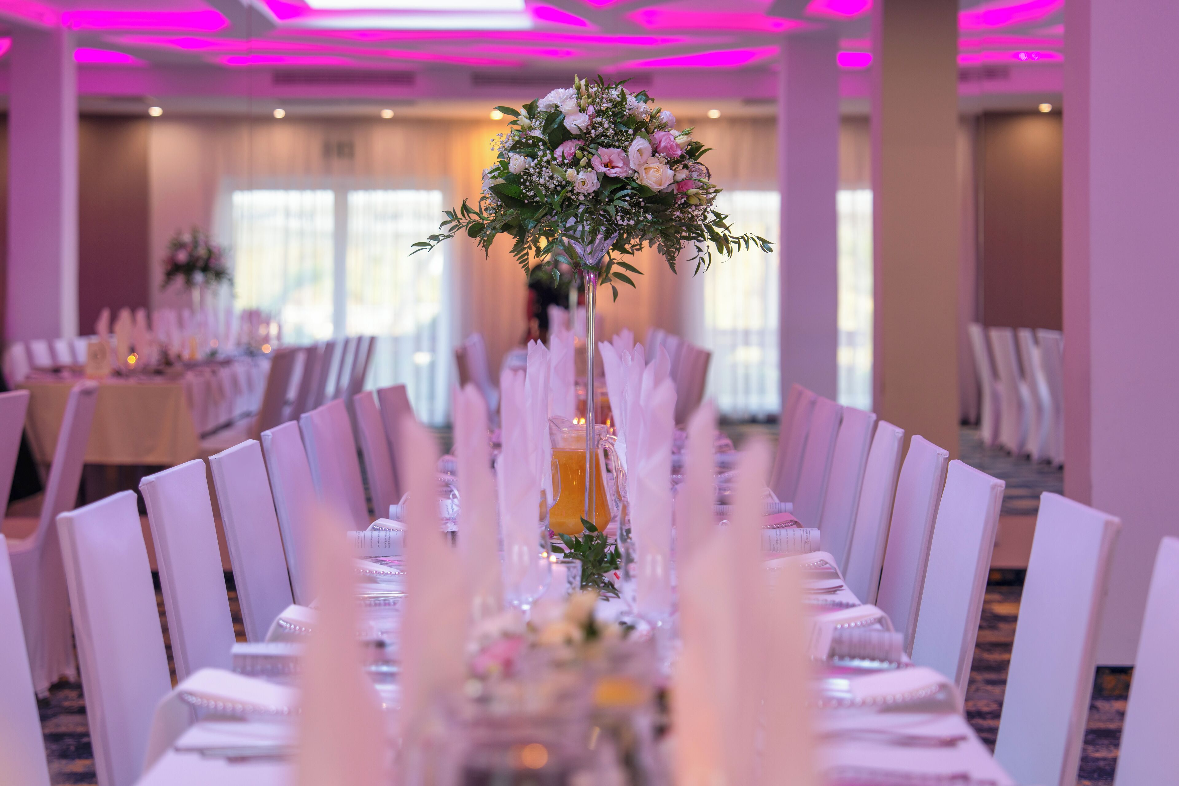 A banquet hall with long tables set for a formal event, featuring a central floral arrangement and a pitcher of orange juice.