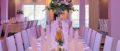 A banquet hall with long tables set for a formal event, featuring a central floral arrangement and a pitcher of orange juice.