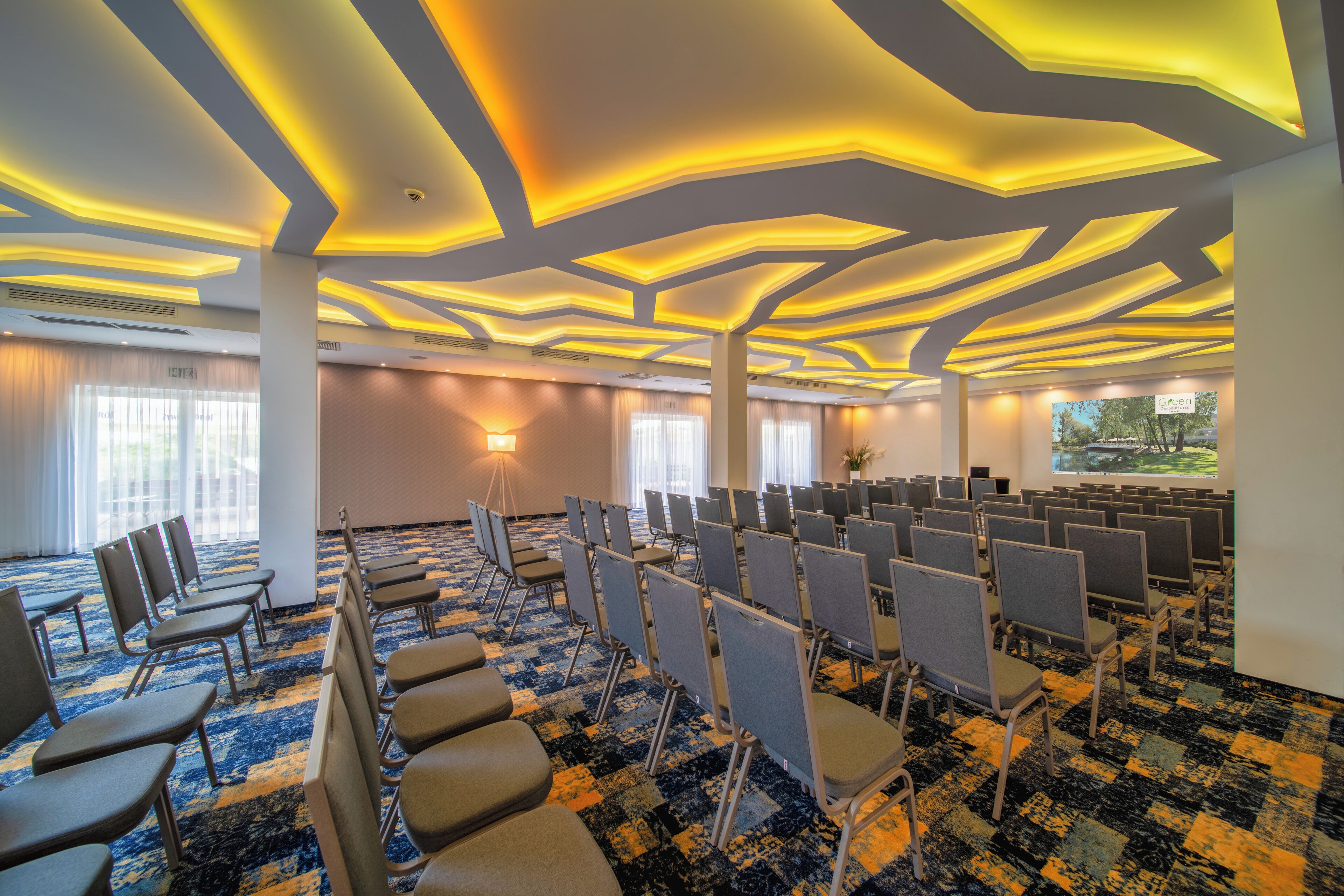 A conference room with rows of chairs, a patterned carpet, and a ceiling with geometric lighting.