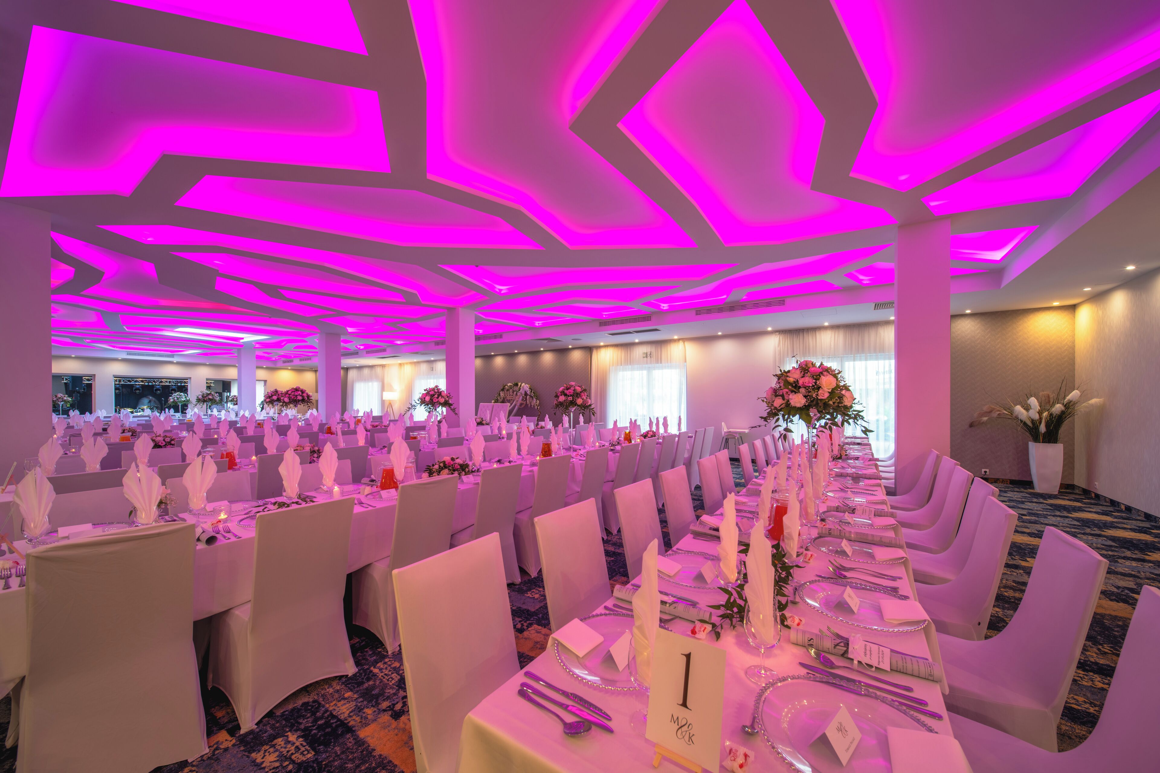 A banquet hall with rows of tables set for a formal event, featuring a modern ceiling design and ambient lighting.