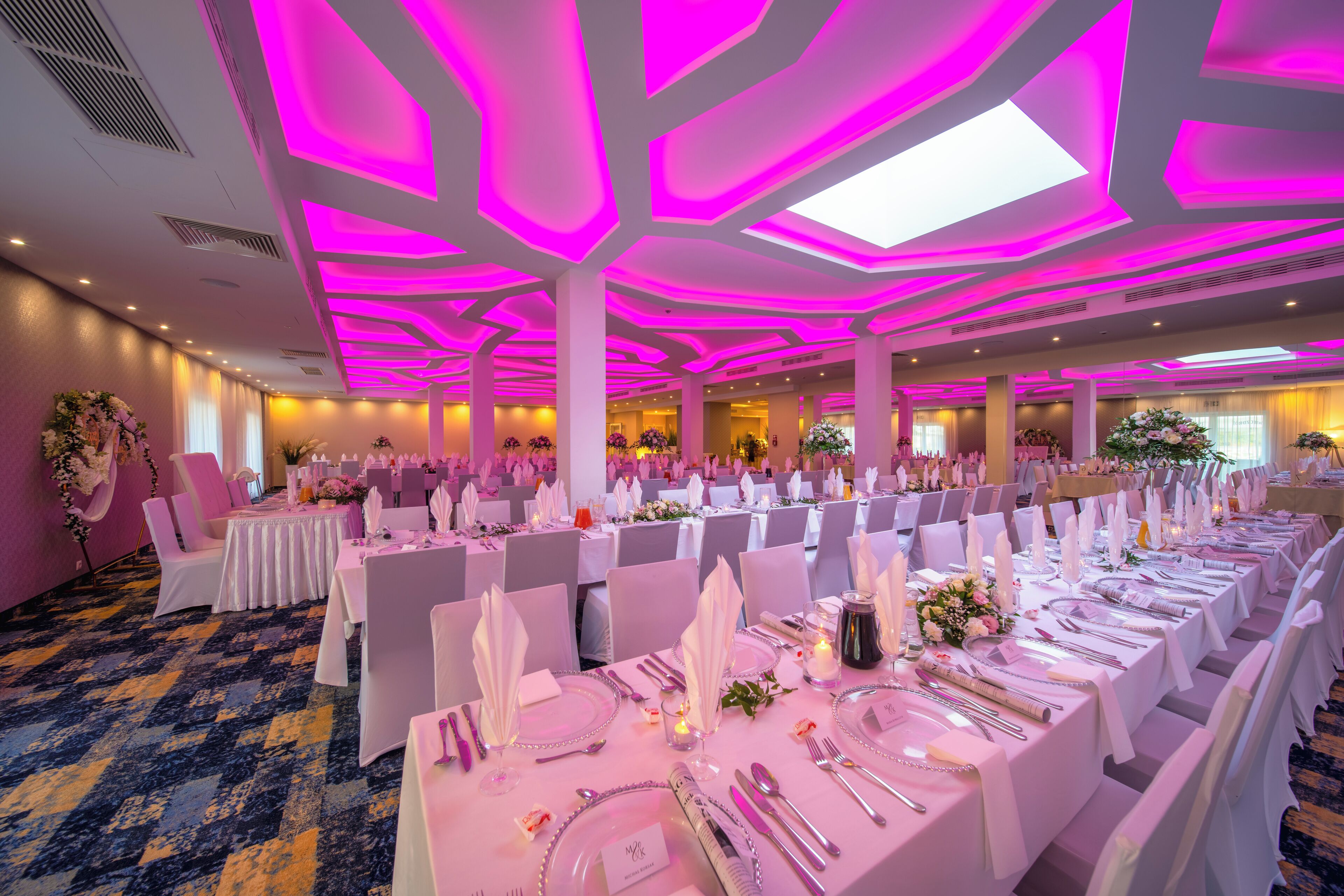 A banquet hall with tables set for a formal event, featuring a ceiling with geometric lighting and a carpeted floor.