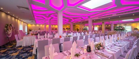Un salón de banquetes con mesas dispuestas para un evento formal, con un techo de iluminación geométrica y suelo alfombrado.