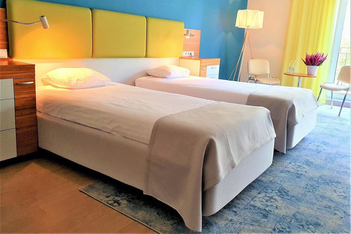 A hotel room with two beds, a wooden nightstand, a desk, a chair, a small table with a lamp, and a potted plant.