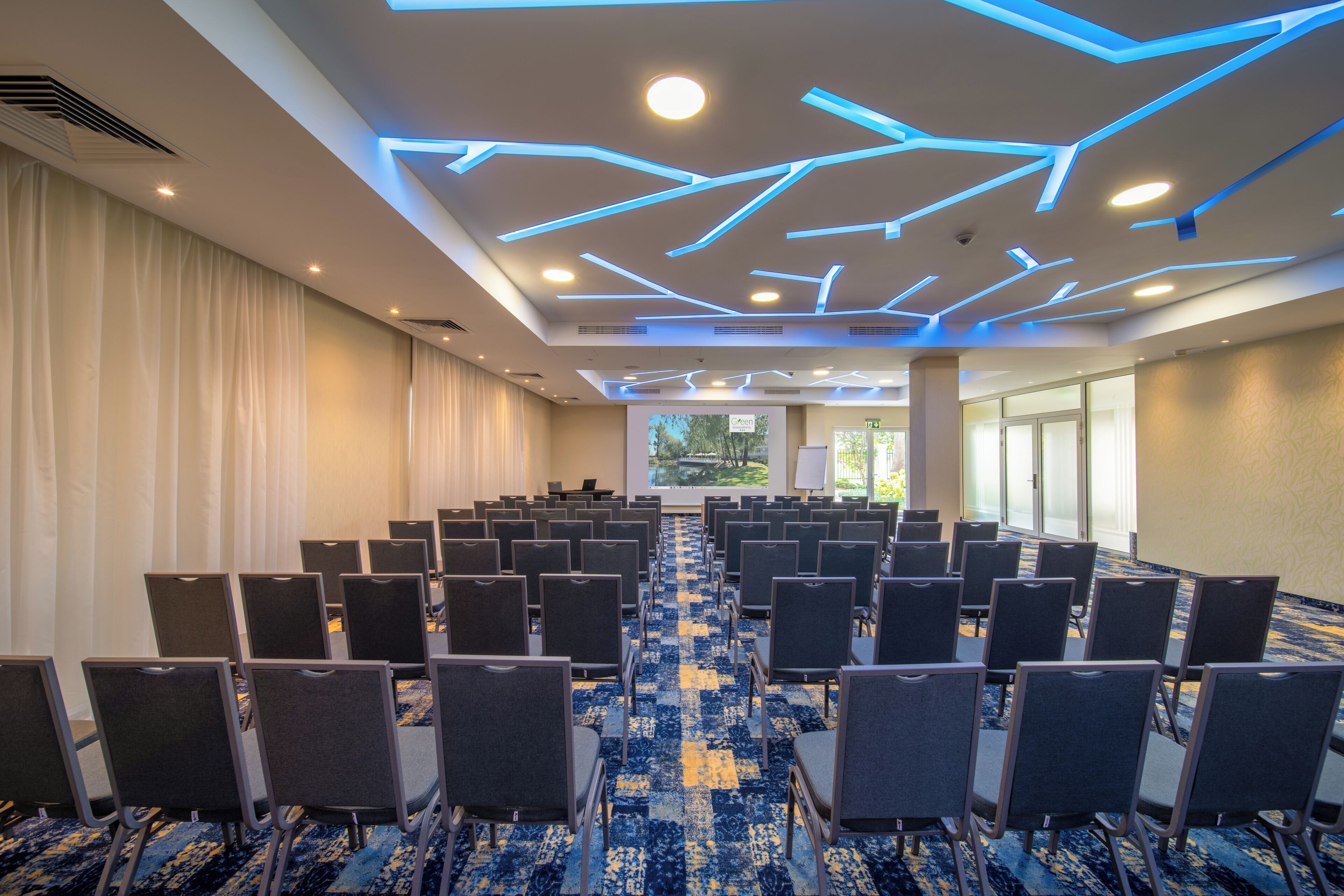A conference room with rows of chairs, a patterned carpet, and a ceiling with geometric lighting.