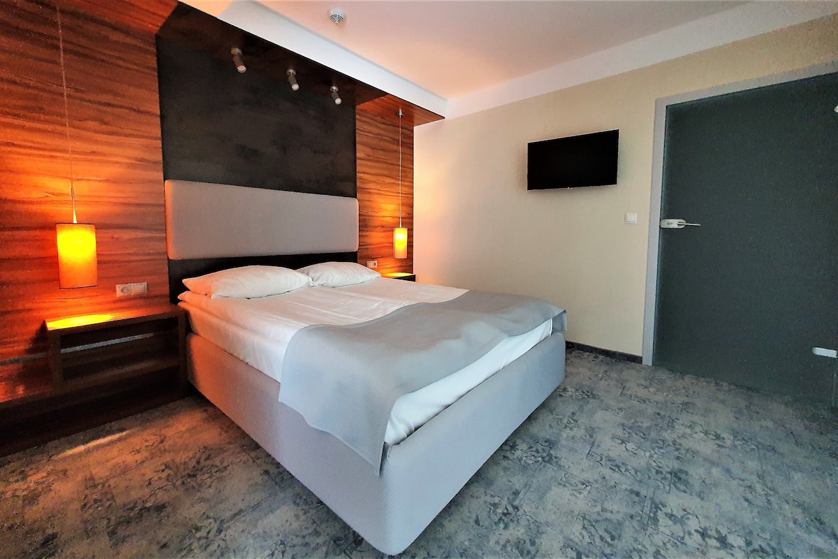 A modern hotel room with a large bed, wooden headboard, two bedside lamps, a flat-screen TV, and a door.