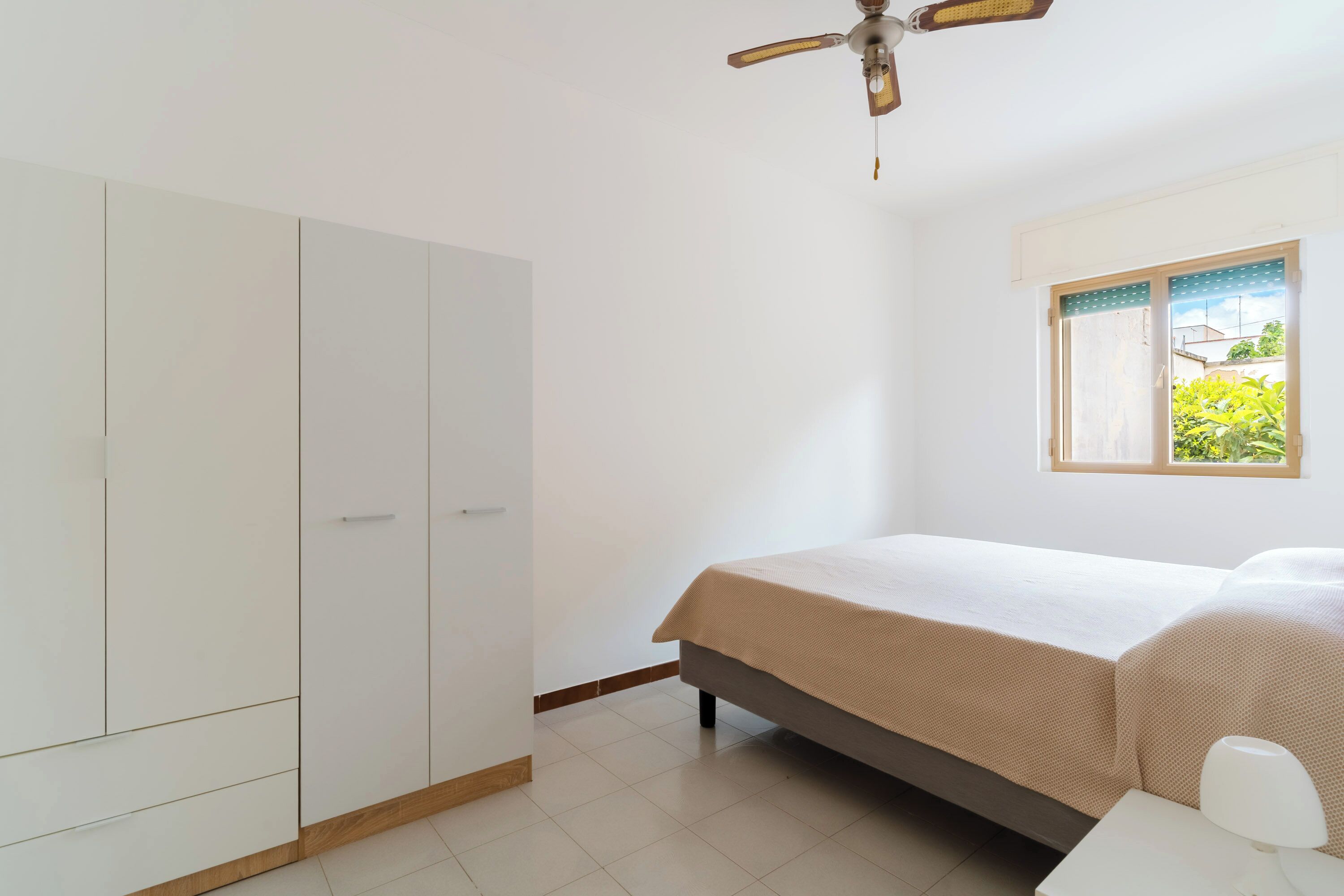 A bedroom with a bed, a wardrobe, a ceiling fan, and a window with a view of greenery.