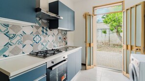 Cocina moderna con estufa a gas, horno y armarios empotrados. Hay una puerta corrediza de vidrio que da a un patio con un árbol.