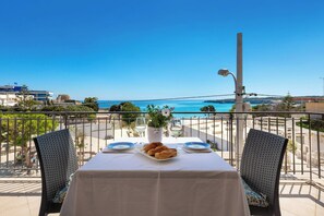 A balcony table with a white tablecloth, two chairs, a plate of croissants, a vase with flowers, and a clear view of the sea and buildings in the background.