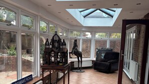 A conservatory with a view of a brick wall and garden, a wooden table with a birdcage, and a sign reading "EDGWAR ROAD".