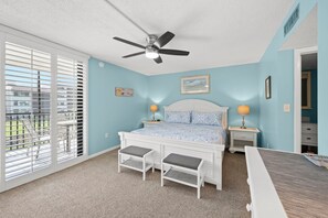A bedroom with a bed, two bedside tables, a ceiling fan, and a view of the outdoors through large windows.