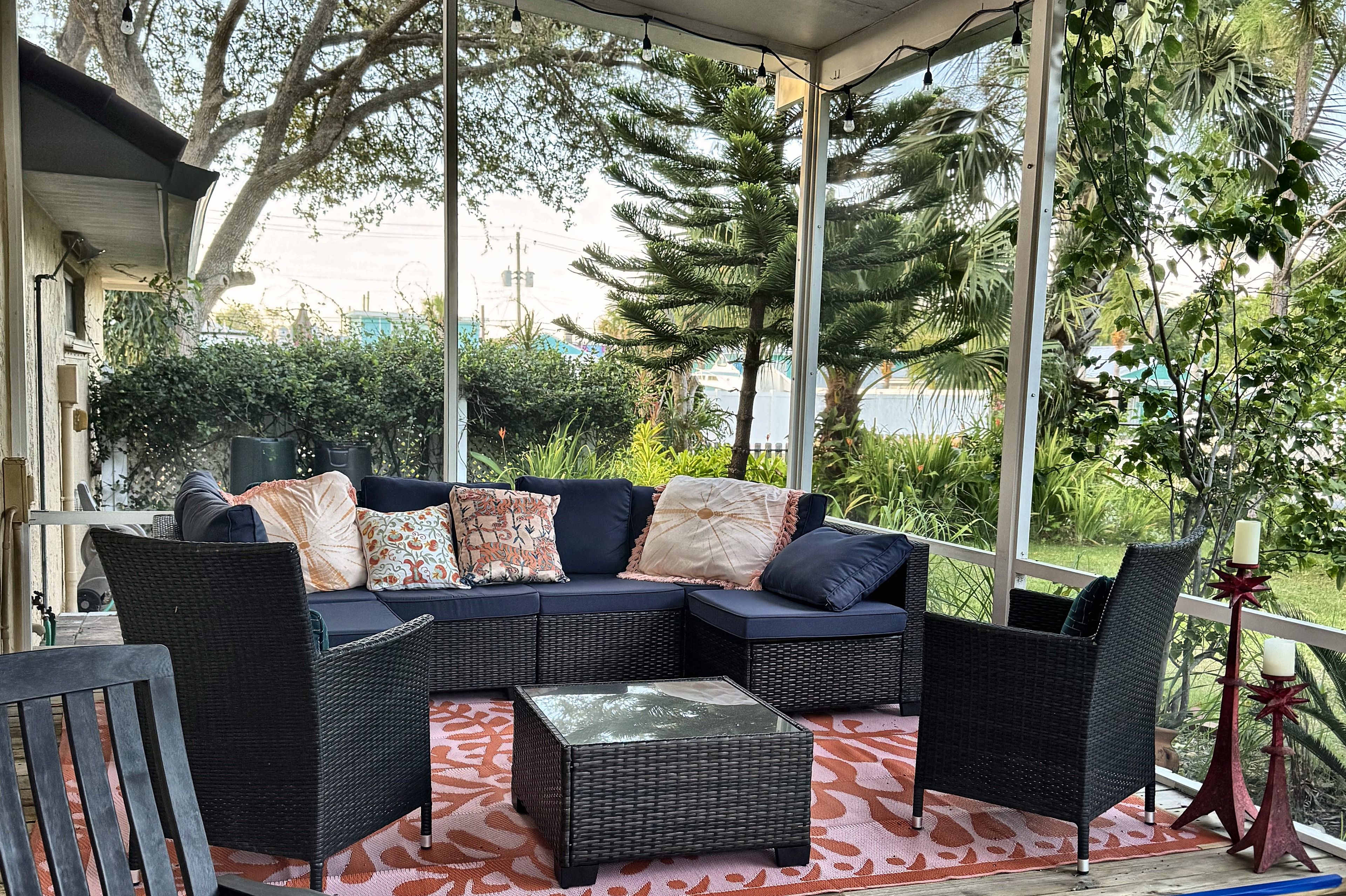 A covered outdoor seating area with a dark-colored sofa, multiple cushions, and a small table.