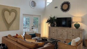 A spacious living room with a large heart-shaped wall art, a flat-screen TV, a wooden cabinet, and a comfortable seating area with a sofa and chairs. - Walk to Beach, Dining, Shops, Screen Patio, Game Room, Hobe Sound Zeus Park (Hobe Sound)