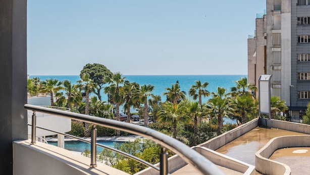 A balcony with a view of the sea, palm trees, and a modern building.
