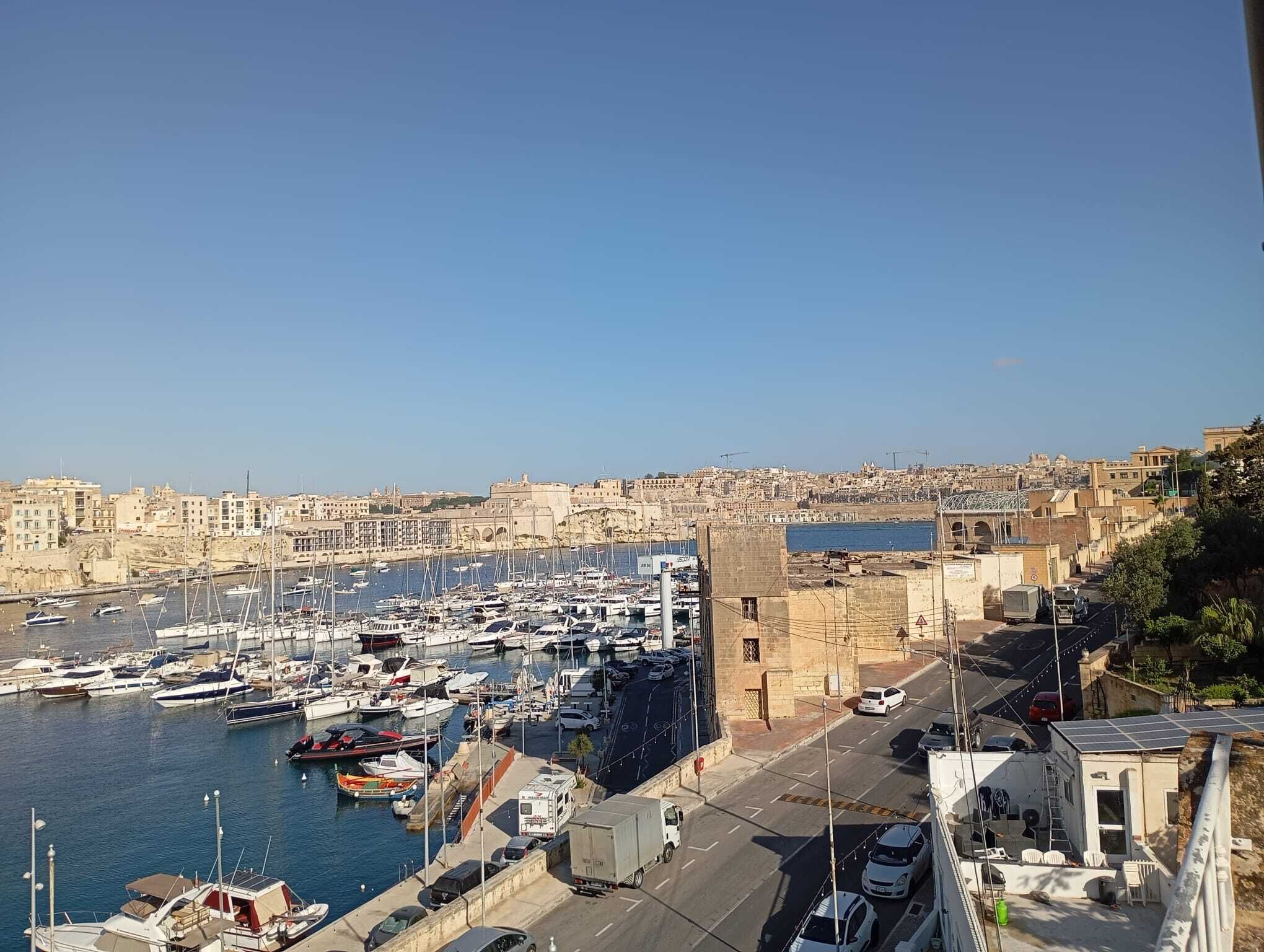 Una marina con barcos amarrados, una carretera con vehículos y edificios al fondo.