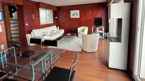 A modern living room with a glass dining table, white sofa, and a white armchair. A flat-screen TV is mounted on the wall, and a refrigerator is visible in the background.