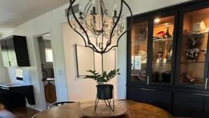 Un comedor con una mesa redonda de madera, sillas blancas y mullidas, una araña y un aparador con puertas de cristal.