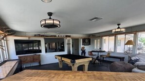 Una habitación amplia con mesa de billar, una mesa de madera con objetos negros y un gran ventanal que proporciona luz natural.