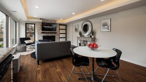 A modern living room with a fireplace, a flat-screen TV mounted on a stone wall, a grey sofa, a dining table with two chairs, and a large window providing natural light. - Bassett Creek Retreat (Golden Valley)