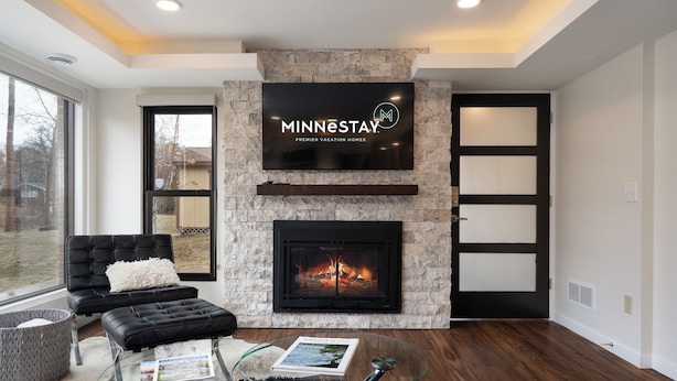 A modern living room with a stone fireplace, a flat-screen TV mounted above it, and a glass coffee table with magazines. - Bassett Creek Retreat (Golden Valley)