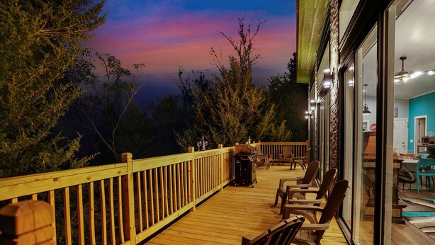 Una terraza de madera con sillas y una mesa, con vistas a un área boscosa y un atardecer.