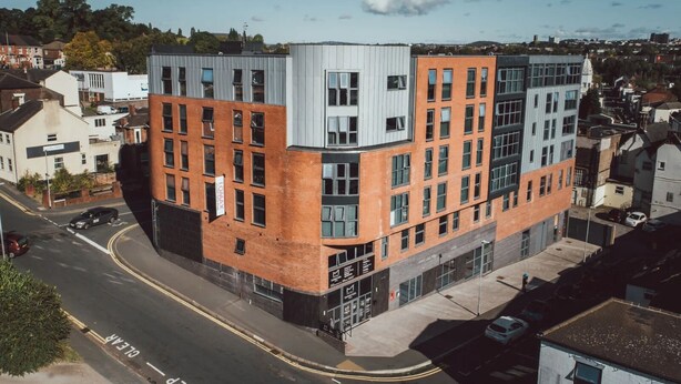 A modern multi-story building with a mix of brick and metal facades, located in an urban setting with visible roads and other buildings. - Student Only Lomax Halls (Stoke-on-Trent)
