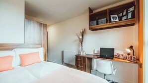 A hotel room with a bed, a desk with a laptop, a radiator, and a shelf with framed photos. - Student Only Lomax Halls (Stoke-on-Trent)