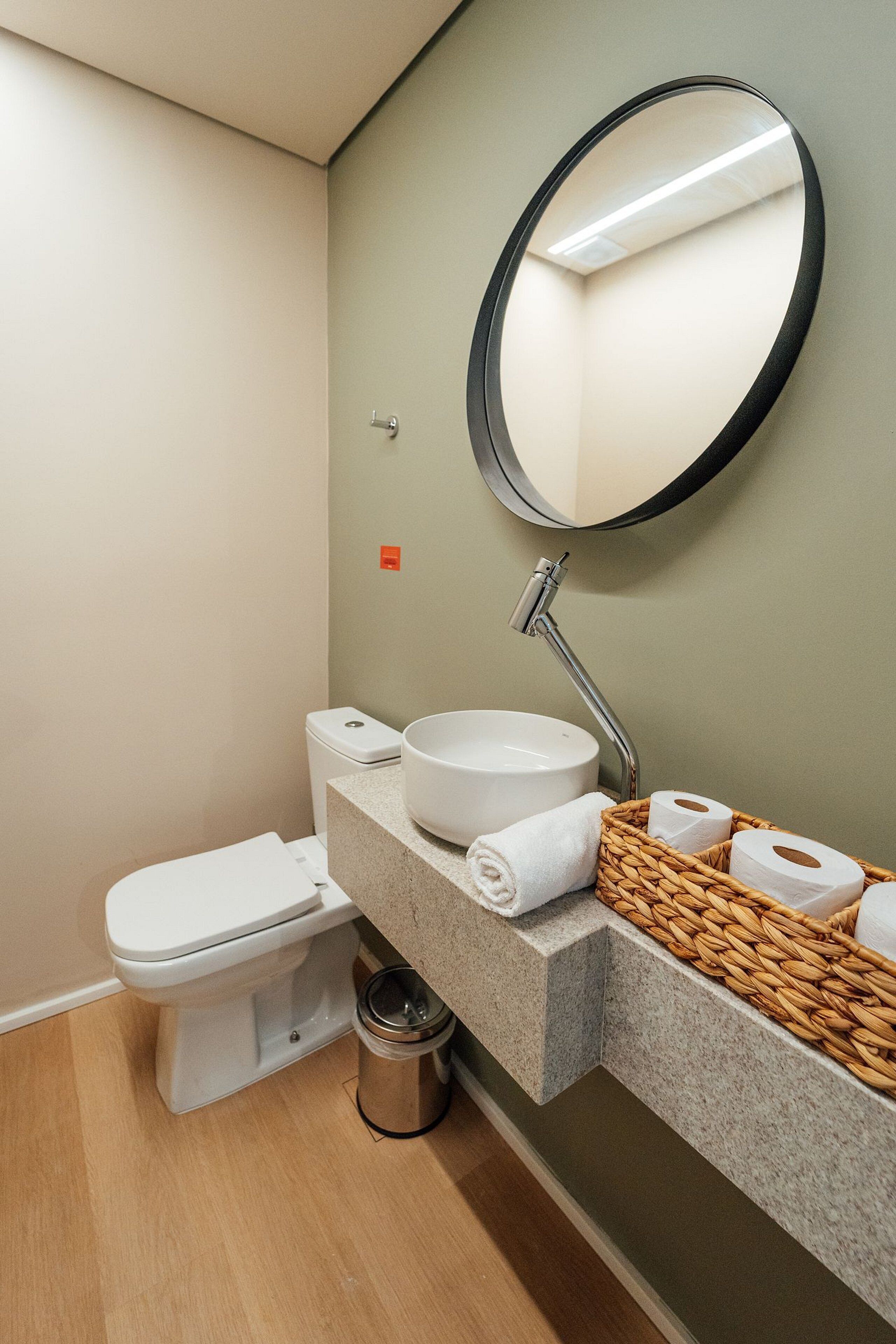 A modern bathroom with a white toilet, a round mirror, a sink with a granite countertop, and a basket of toiletries.
