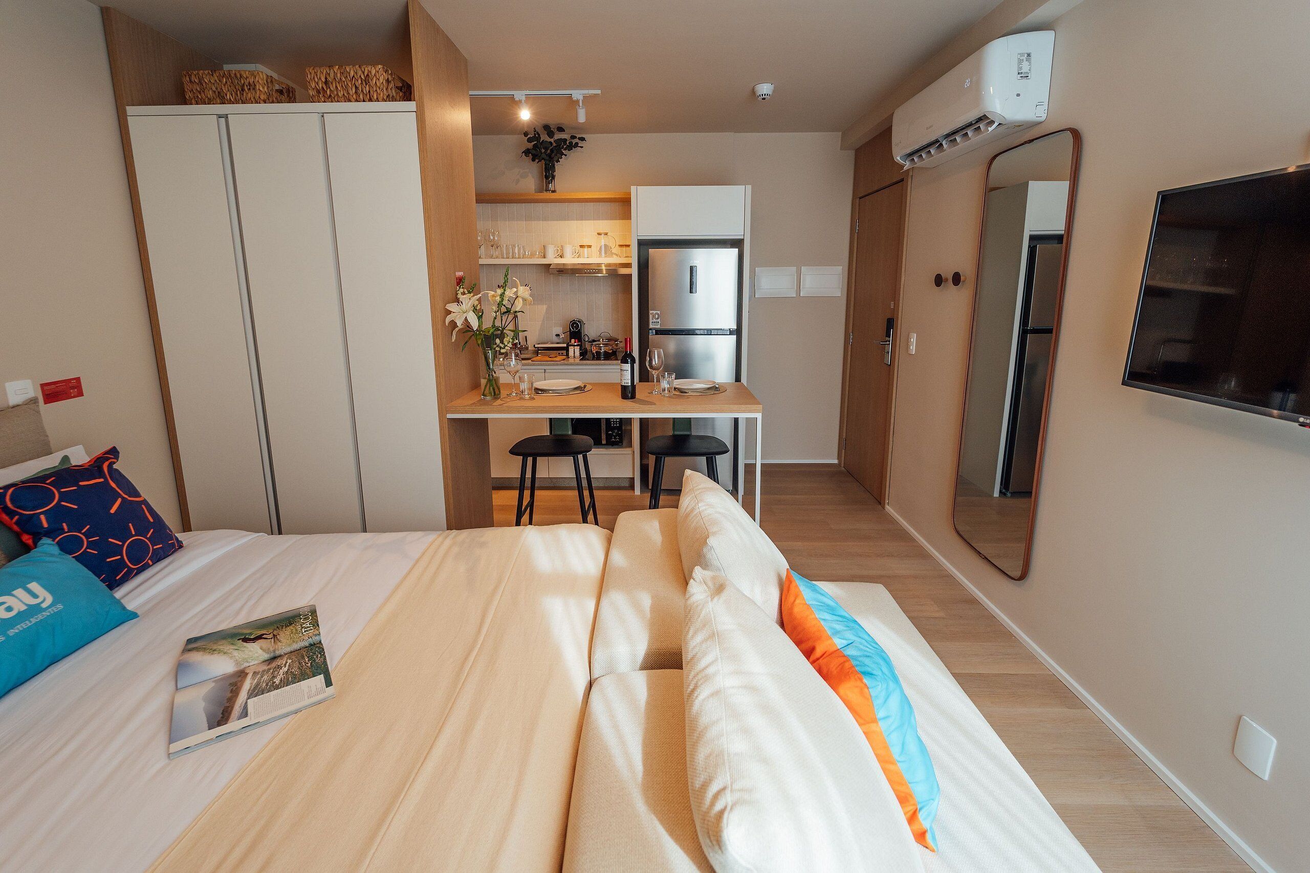 A compact hotel room with a bed, a small kitchenette, a dining area, and a TV mounted on the wall.